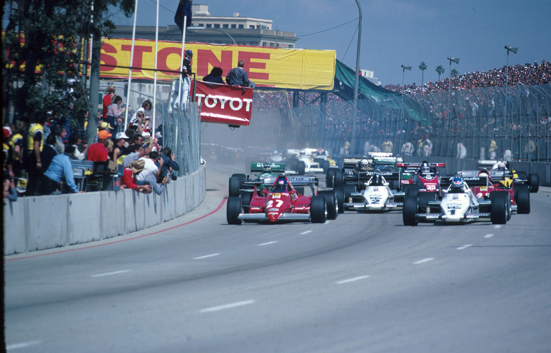 Several racecars drive down a track with onlooking fans in the stands.