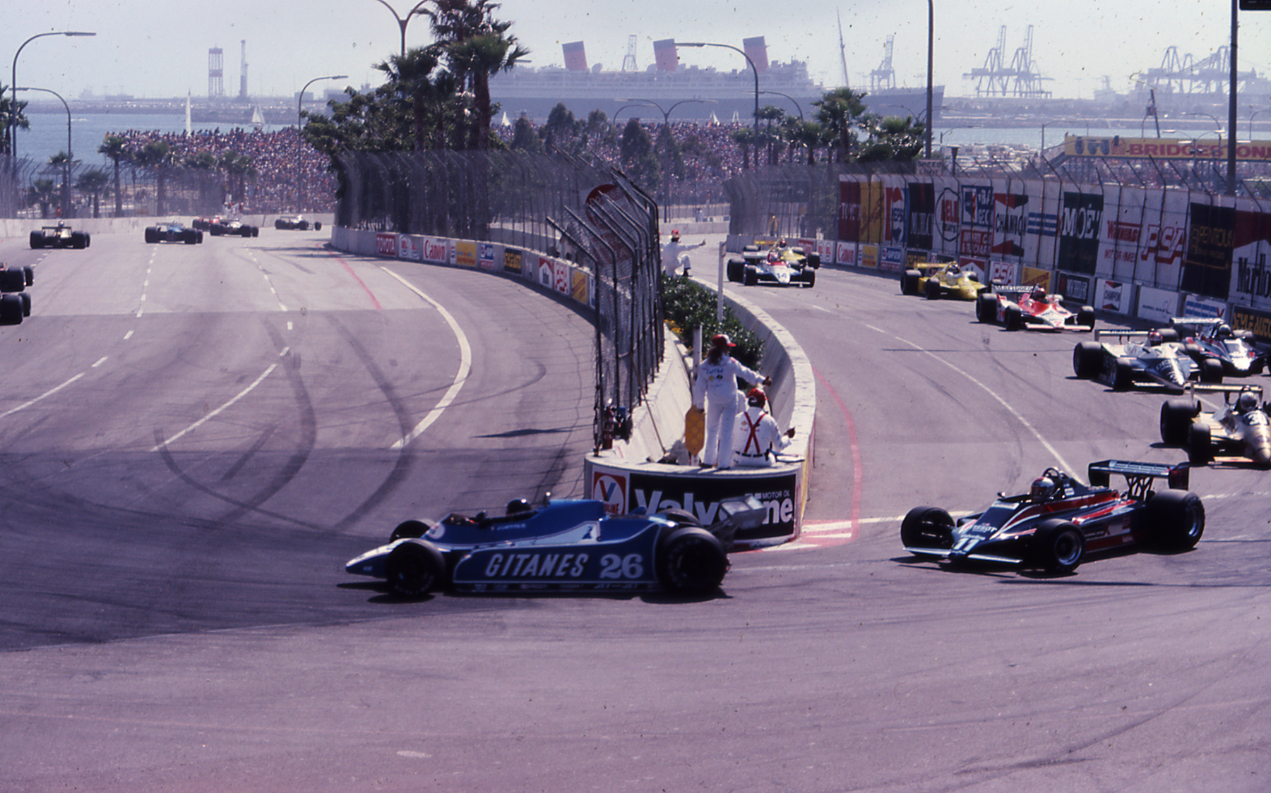 Racecars prepare for a hairpin turn on a track with the Queen Mary in the distance.