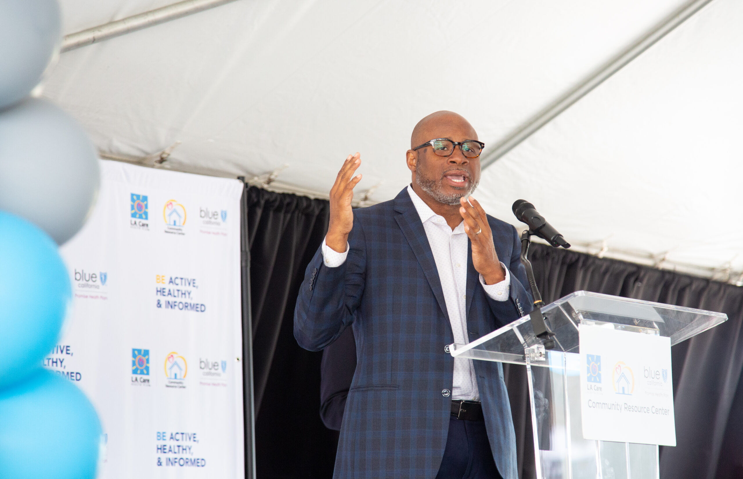 Councilmember Al Austin speaks into a podium. Balloons are to his left, and the background has various Blue Shield and L.A. Care logos.