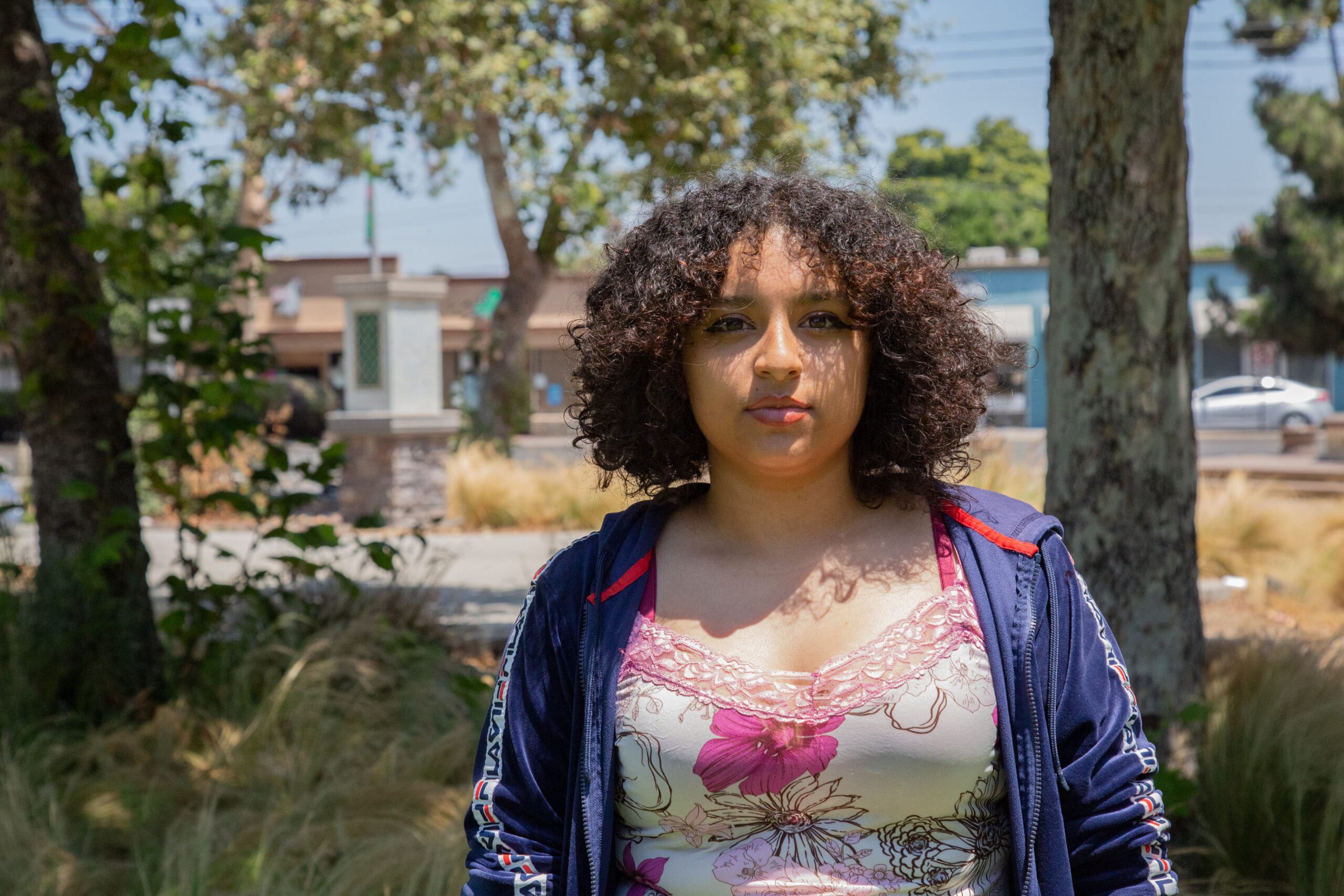 A girl wearing a white shirt with a pink flower print, and a blue jacket stands in the shadows. Buildings and trees are behind her and out of focus.