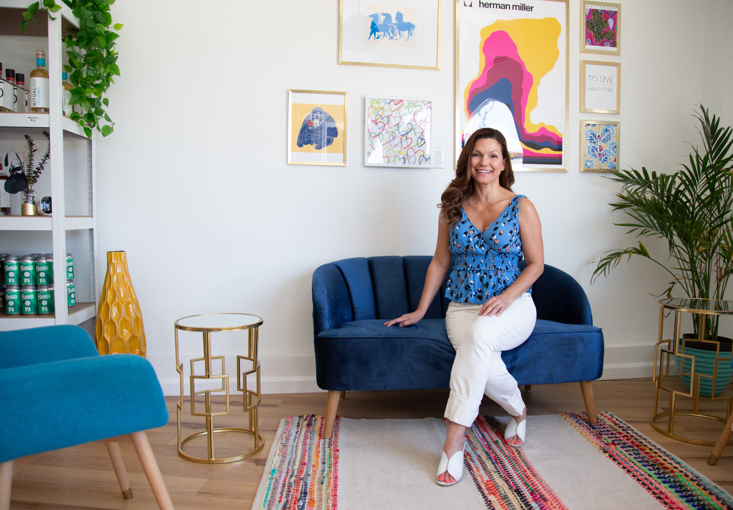 A woman with a blue shirt and white pants sits on a blue couch. Art is on the wall behind her.